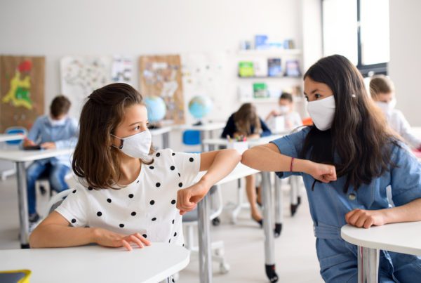 Two girls wearing masks sitting in class and bumping elbows