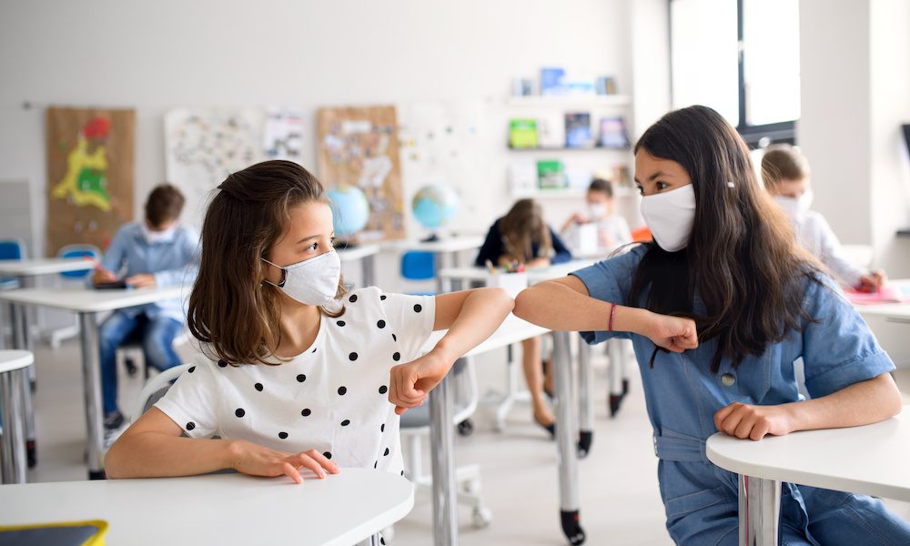 Two girls wearing masks sitting in class and bumping elbows
